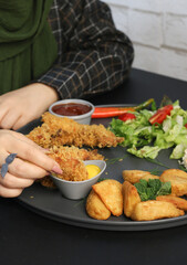 Hand dipping crispy fried shrimp into mustard sauce, served with potato wedges and salad on a dark plate, perfect for fast food advertising, seafood restaurant menus, and casual dining promotions.