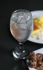 Close-up of a frosted glass filled with crystal-clear ice cubes, refreshing cold water drink placed on a dark table beside a dish of grilled meat and fluffy saffron rice, bright natural lighting creat