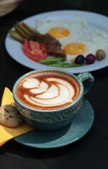 Artistic cappuccino coffee with beautiful foam pattern placed beside a plate of fried eggs, grilled meat, pickles, tomatoes, and olives on a rustic table, highlighting a comforting countryside