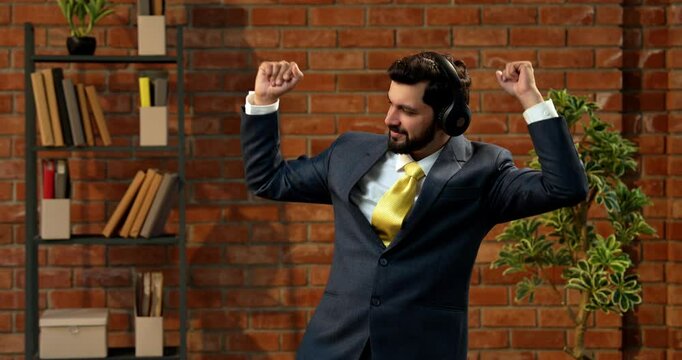 Dancing joyfully while wearing headphones, Indian businessman enjoys upbeat music break in modern office, smiling and moving in rhythm against cozy terracotta brick wall in formal attire