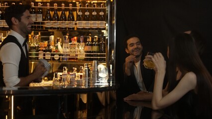 Smart bartender serving gin fizz or tonic with sliced lemon at luxurious bar, engaging lively conversation with VIP customer couple at side view in atmosphere at lighting warm nightclub. Vinosity.