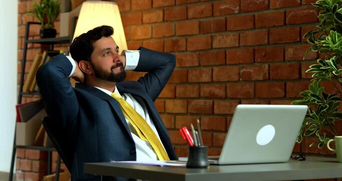 Taking a break while working, cheerful Indian businessman pauses and stretches arms after long laptop session at desk in cozy modern office with terracotta brick wall, smiling in formal wear