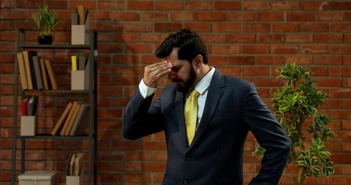 Stressed or worried Indian Asian young businessman or CEO with beard in formal suit standing alone in cozy modern office interior with terracotta brick wall background looking tense and anxious