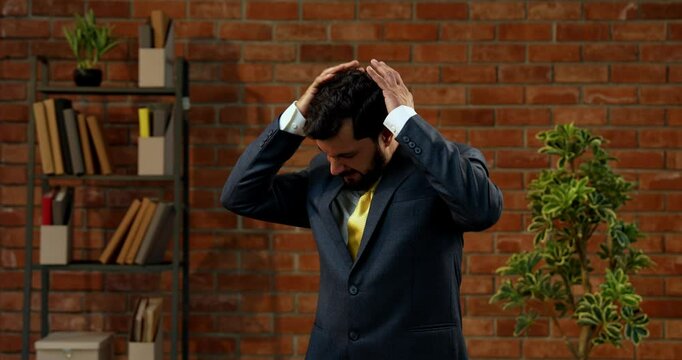 Stressed or worried Indian Asian young businessman or CEO with beard in formal suit standing alone in cozy modern office interior with terracotta brick wall background looking tense and anxious