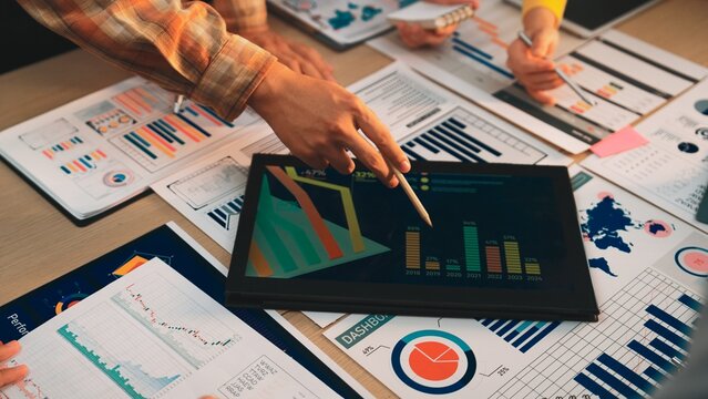 A diverse business team engages in collaborative data analysis using charts and graphs displayed on a tablet, surrounded by reports in a modern office setting. SACTR