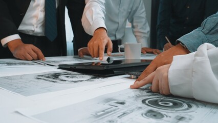 A dynamic office meeting scene shows a diverse team engaged in analysis and collaboration, exploring blueprints and discussing innovative ideas around a laptop. SACTR