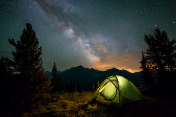 Illuminated green tent nestled in mountain landscape under a night sky filled with stars and the Milky Way, showcasing adventure and serenity