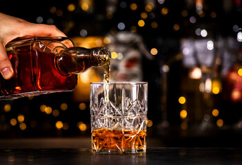 Bartender pours bourbon or whiskey in glass. Black bar counter background with hard drinks bottles...