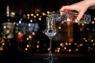 Bartender pours Italian vodka grappa in glass. Bottles of strong drinks on black bar counter background with lights