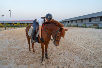 Front view of teen girl stay on the horse and bend down to make horse to relax before train or practice in the arena.