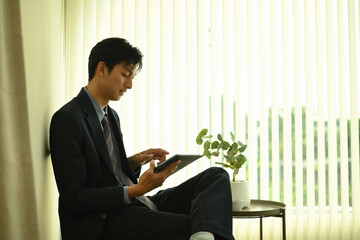 A young businessman sits near a large window with vertical blinds, using a digital tablet in a bright, minimalist office space