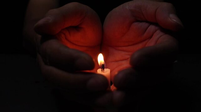 Hands Protecting a Burning Candle from wind in a Dark and Peaceful Setting. The candle flame illuminates the palms, symbolizing the bitterness of loss, death, and grief.