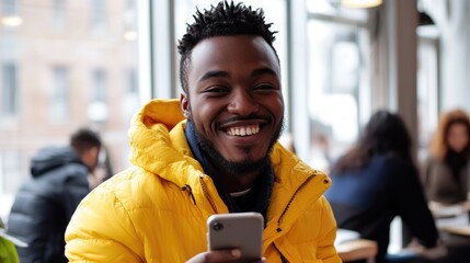 A smiling man with a beard stands in a modern office environment, wearing a tan jacket over a white shirt, holding a smartphone in his hands.