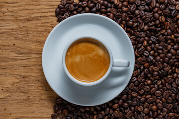 Fresh brewed espresso in a white cup on a saucer and surrounded by roasted coffee beans on a rustic wooden table, closeup, top view. Concepts of coffee culture, caffeine, breakfast and relaxation