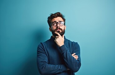 Young man wears sweater, glasses over blue background. Handsome guy with beard looking up thoughtfully. Facial expression indicates thought worry. Portrait of man with glasses against blue background.