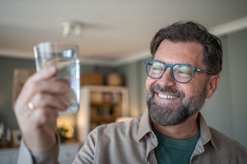 Happy mature man holding and observing glass of water at home