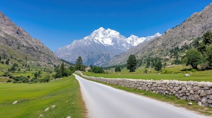 Naklejka premium Mountain road through a valley. Scenic paved road winds through a lush green valley, leading to a majestic snow-capped mountain peak. A stone wall borders the road, with fields of grass and trees