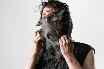 A curly-haired guy in a grunge outfit pulls a tattered rag off his face, hiding his face from...
