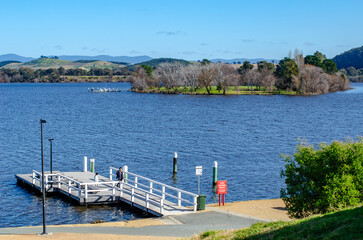 A pier or small dock extending into Lake Burley Griffin, with a wooden walkway and railings leading to the water surrounded by rolling hills and mountains in Canberra, the capital of Australia.