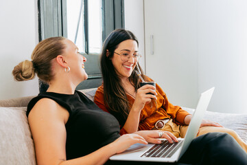 Two Latin young women, coworkers, relax on the couch working remotely and sharing mate