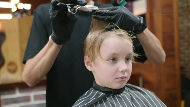 Cute school age boy getting haircut in barbershop. Children hairdresser with professional tools - comb and scissors. Cutting hair for kids. Services of a professional stylist.