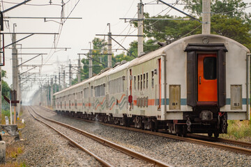 Naklejka premium Passenger train of PT KAI moves through West Java's railway network, framed by overhead wires and greenery on both sides.