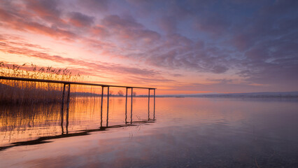 Fototapeta premium A serene sunrise over Lake Constance with a wooden pier extending into the calm water. The sky transitions from soft pink to deep blue, reflecting on the lake’s surface.