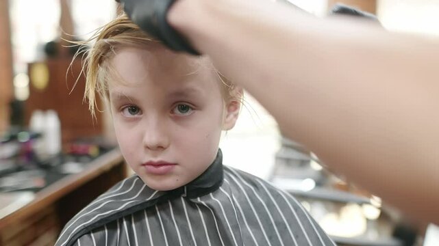 Cute school age boy getting haircut in barbershop. Children hairdresser with professional tools - comb and scissors. Cutting hair for kids. Services of a professional stylist.