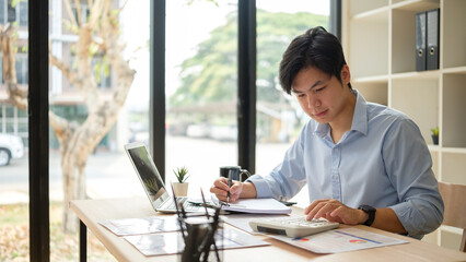 Young businessman calculating budget and reviewing financial reports at desk in modern office.