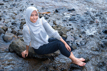Asian Muslim women sit on a stone in the river, a woman sitting on a riverbank with a flow of water flowing between the rocks.