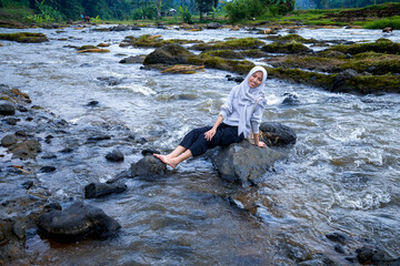Asian Muslim women sit on a stone in the river, a woman sitting on a riverbank with a flow of water flowing between the rocks.