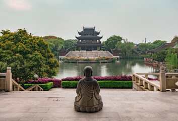 Serene view of a stone Buddha facing a traditional pavilion near a tranquil lake in a Chinese garden