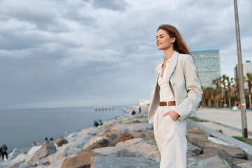 Young woman in beige pantsuit enjoying calm seaside breeze with modern city and cloudy sky...