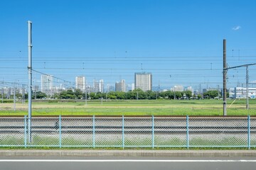 Fototapeta premium Urban landscape, rail tracks and open space. A view of a city skyline behind a grassy field and metal fence, with train tracks in the foreground. Clear, blue sky