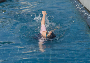 Asian Young girl practicing swimming strokes in clear blue water during sunny day, wearing goggles and swimwear, showcasing active lifestyle and dedication