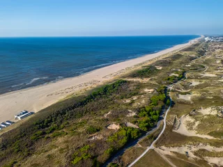 A stunning aerial view of a long, sandy beach stretching along the coast, bordered by dunes and greenery, with a clear blue sky and calm sea creating a peaceful, natural atmosphere.
