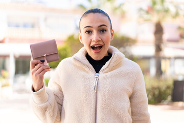 Young moroccan girl holding a wallet at outdoors with surprise and shocked facial expression