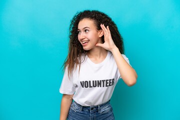 Young Arab volunteer woman isolated on blue background listening to something by putting hand on...