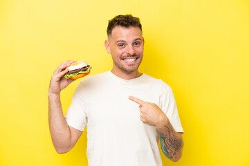 Young caucasian man holding a burger isolated on yellow background with surprise facial expression