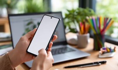 Person holding a smartphone with blank screen,  in front of laptop and colored pencils