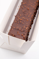 A rectangular chocolate eclair, generously coated in a glossy dark chocolate glaze and sprinkled with chopped nuts or crunchy elements, rests inside a white paper tray.