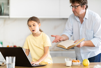 Fototapeta premium Father invites his little daughter to read an interesting book together. Daughter refuses and continues to play games on laptop