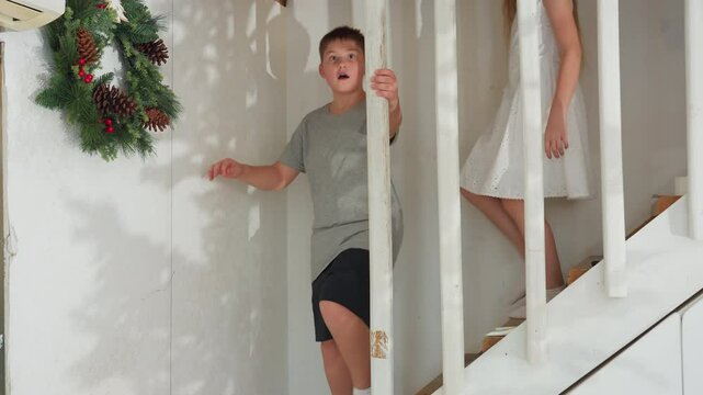 kids in white socks, boy in gray top and black shorts, girl in white gown coming down staircase, boy with surprised expression, girl smiling, playful indoor family moment, festive holiday decorations