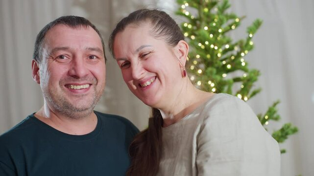 Portrait view of happy husband and wife smiling warmly at each other while standing together in cozy festive room decorated with Christmas tree and glowing lights, expressing love, connection