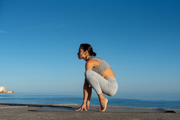 Young woman practicing forward bend yoga pose outdoors on a sunny day by the sea, wearing gray fitness attire, promoting health, flexibility, and mindfulness