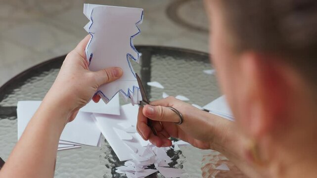 side view of woman carefully cutting white folded paper with blue outlined shapes using scissors on glass table during creative crafting session with small paper pieces scattered around surface