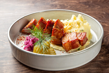 A delicious plate featuring glazed pork chunks served with creamy mashed potatoes, garnished with dill and accompanied by pickled cabbage and vegetables, creating an appetizing presentation.
