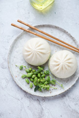 Plate with chinese steamed baozi buns and edamame beans on a white stone background, vertical shot, high angle view
