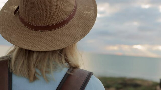Female traveler wear backpack standing on coastal cliff, gazing across expansive ocean landscape with dramatic cloudy horizon during golden evening light.Europe travel. Lifestyle, vacation, tourism,