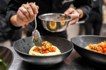 A chef artistically plating a dish of hummus topped with fresh diced tomatoes and herbs. The focused hands and vibrant ingredients highlight the culinary creativity in a professional kitchen.